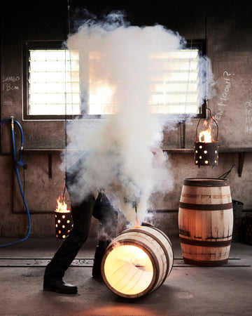 Person in a dark outfit with smoke effects in an industrial setting with barrels and lights.