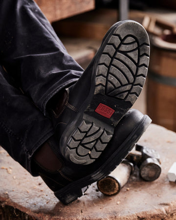 Close-up of a person wearing black work boots with a visible brand logo, standing on a wooden surface.