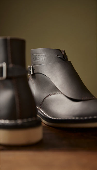 A close-up of a dark brown leather ankle boot with a strap and buckle, featuring a debossed HUGO logo on the side, set against a warm brown background.