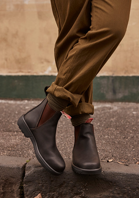 A person wearing dark brown Chelsea boots with brown cuffed trousers, standing on a stone curb outdoors.