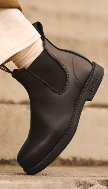A close-up of a person wearing a black Chelsea-style ankle boot with a ribbed elastic side panel and a low chunky heel, paired with a cream ribbed sock and light-colored pants. The brand name 'ROOF' is embossed on the heel.