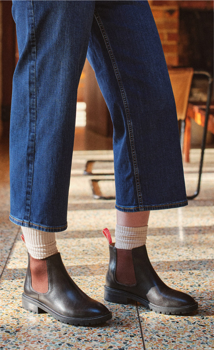 A person wearing dark brown leather Chelsea boots with brown elastic side panels, paired with cream ribbed socks and blue cropped jeans, standing on a speckled terrazzo floor.