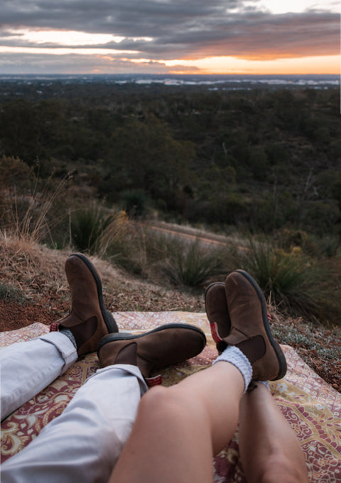 A person reclining outdoors on a patterned blanket at sunset, with their legs raised and wearing brown suede Chelsea-style ankle boots, white pants, and white socks over bare legs, overlooking a forested hillside landscape.